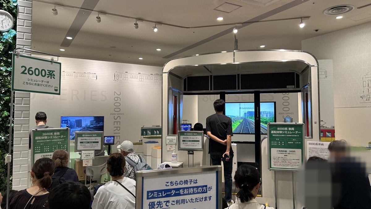 SANZEN-HIROBAの運転シミュレーターの前。8000系の運転台で前の人が運転中で、列に並ぶ人たちが待っている
